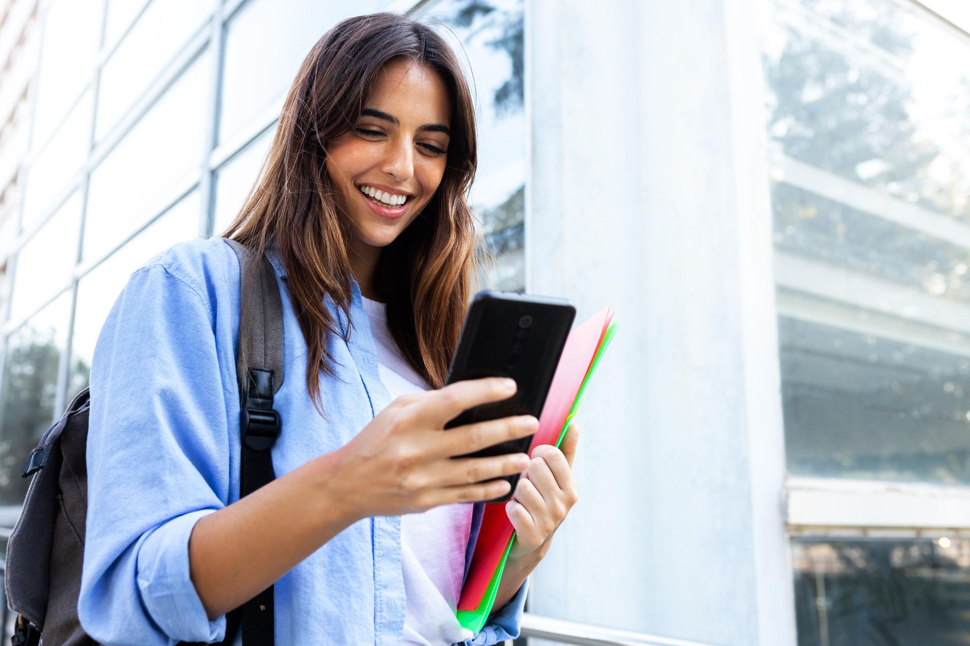 Happy young female university student looking mobile phone standing outside university building. Copy space. Education concept.